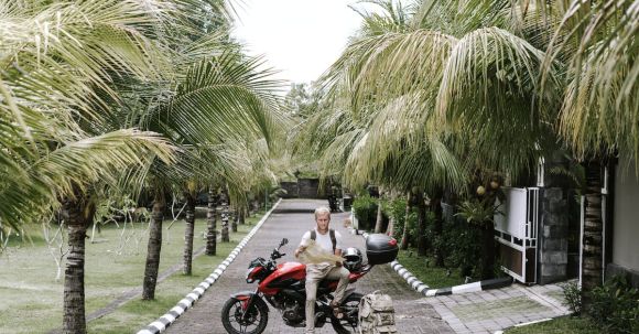 Motorcycle Insurance Guide - Full length male traveler in casual outfit leaning on motorcycle near duffle bag while studying city map on pavement of street among tropical trees in front of open gates of building