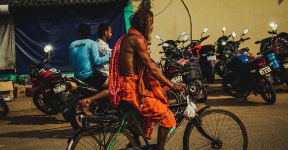 Moto Riding - Side view of barefooted mature Indian male with long hair and beard in traditional clothes riding bicycle on asphalt road near unrecognizable men sitting on motorcycle at night