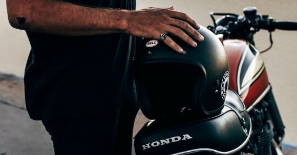 Motorcycle Safety - Side view of anonymous ethnic male biker wearing black clothes standing near modern motorbike with helmet on street near concrete fence