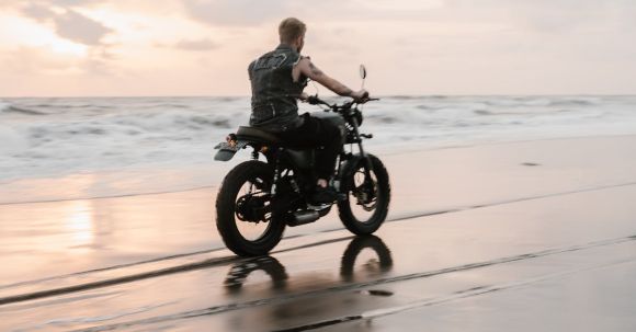 Moto Riding - Back view of anonymous tattooed biker in sleeveless shirt riding motorcycle on wet sand with traces near sea with foamy waves under cloudy sky at sunset