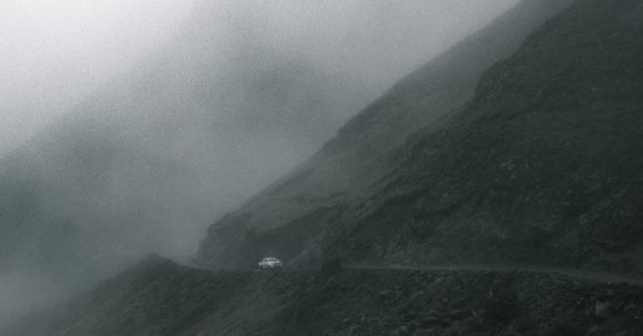 Off-road Maneuvering - Lonely car driving on narrow road in mountainous valley against gray cloudy sky on foggy day