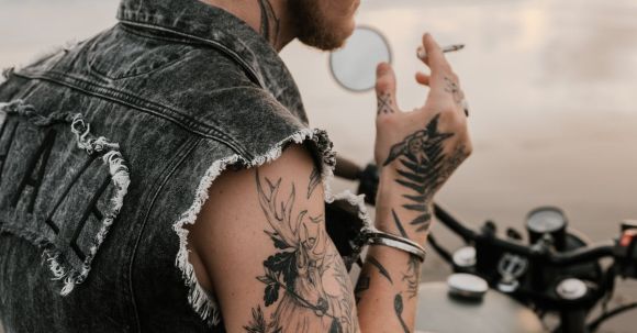 Moto Riding - Side view of thoughtful brutal blond bearded hipster in sunglasses and metal wristband sitting on motorbike while smoking cigarette on sea shore in evening