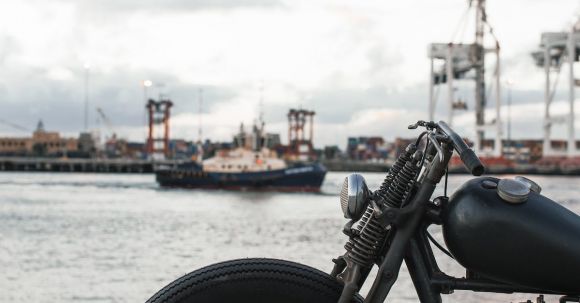 Motorcycle Maintenance - Front part of classical motorcycle with big round wheel and handles parked on pavement next to port for ships under overcast sky