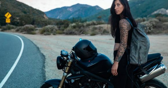 Motorcycle Exploration - Tattooed Woman with Backpack Standing Beside a Black Motorcycle by the Road
